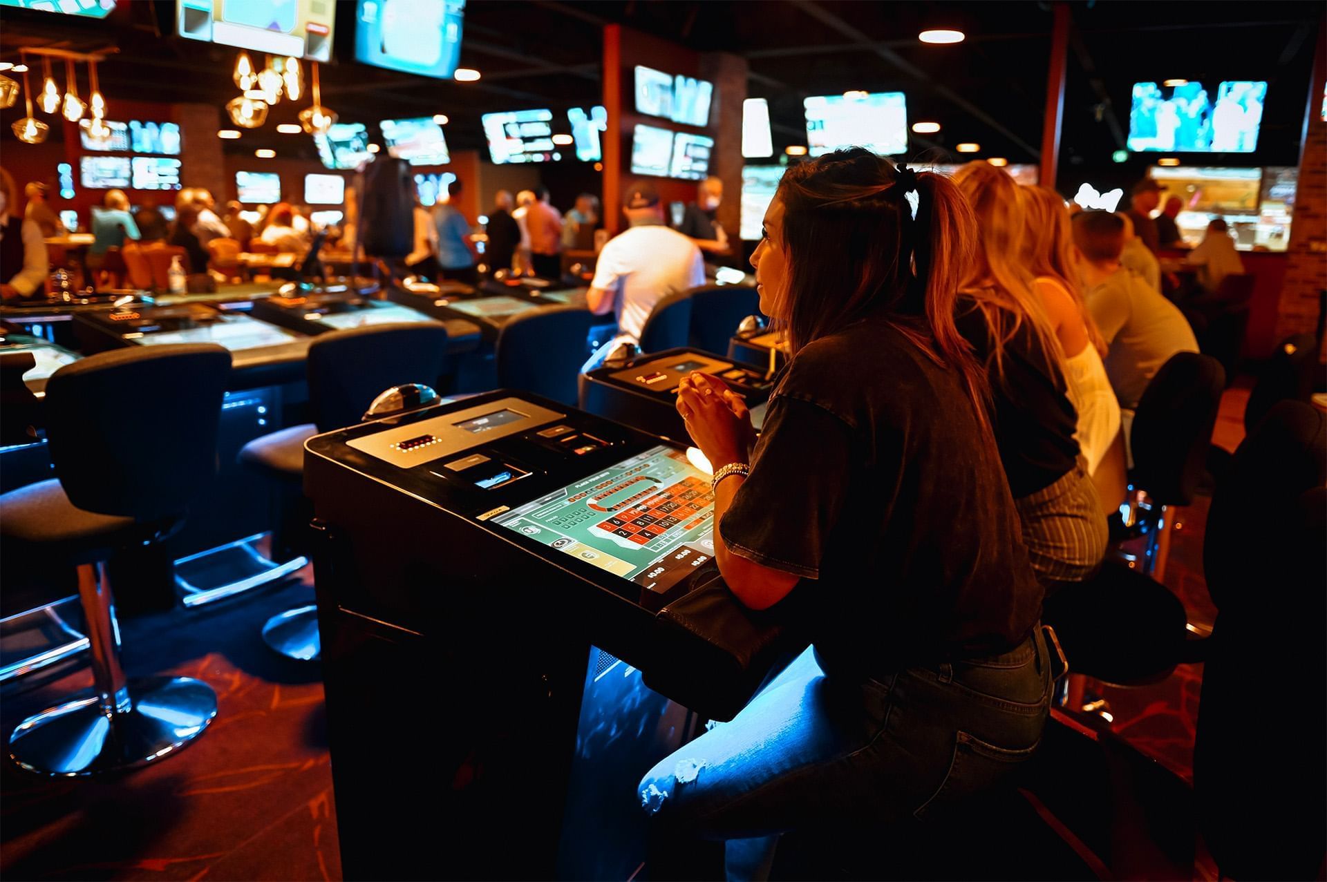 Guests seated at electronic roulette terminals by rows of screens and blue stools under hanging lights at The Brook