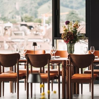 Geräumiges Restaurant mit großen Fenstern, Holzmöbeln, braunen Stühlen und Blumenarrangement auf dem Tisch.