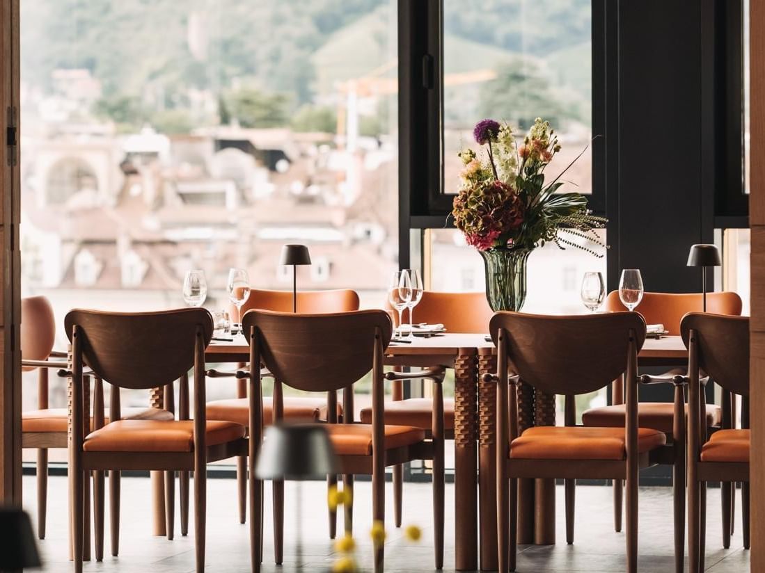 Geräumiges Restaurant mit großen Fenstern, Holzmöbeln, braunen Stühlen und Blumenarrangement auf dem Tisch.