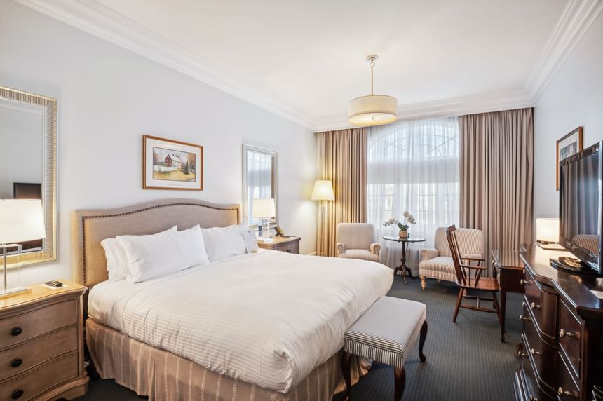 Bed beside work desk, striped bench, and two armchairs near large window in Luxury King at Avon Old Farms Hotel
