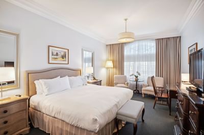 Bed beside work desk, striped bench, and two armchairs near large window in Luxury King at Avon Old Farms Hotel