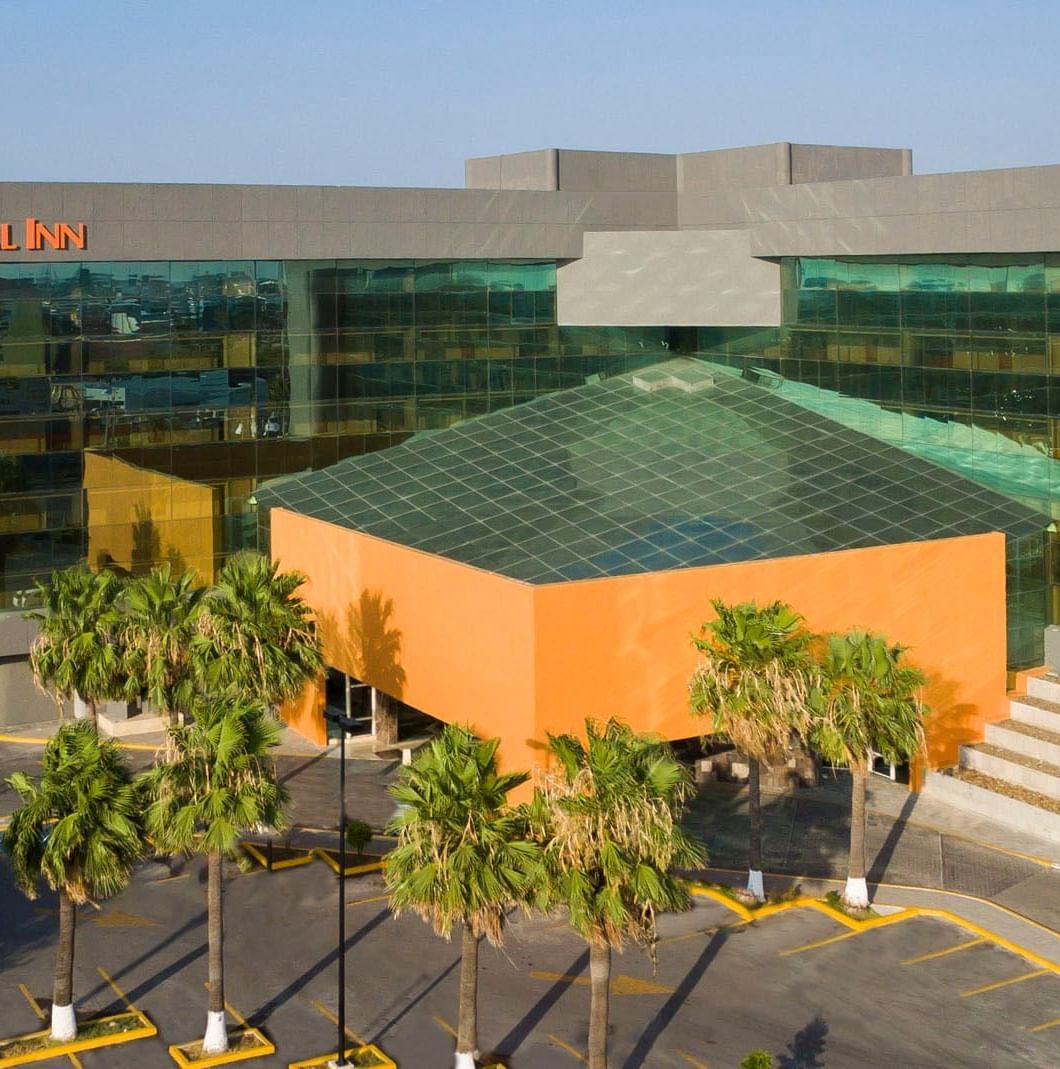 Daytime exterior of Real Inn Nuevo Laredo, showing the modern glass architecture, orange accents, and palm trees