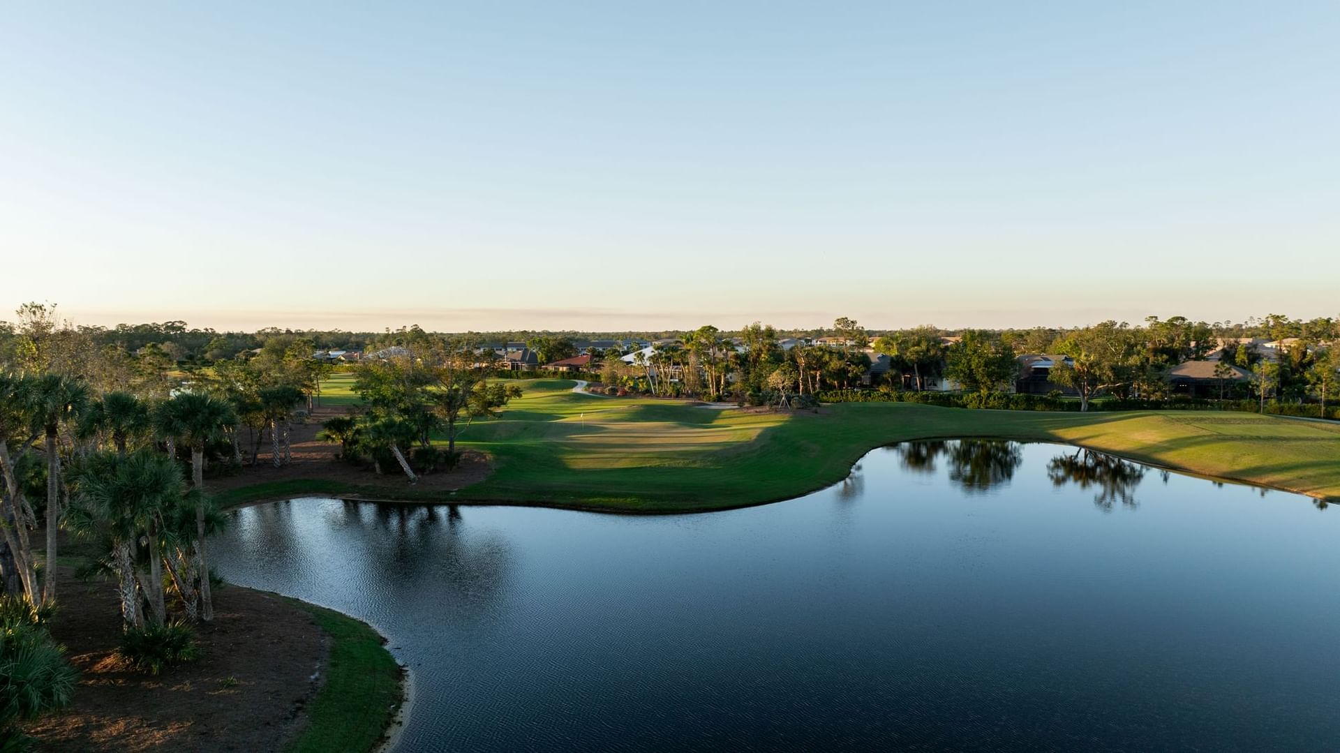 Aerial view of a golf course with a pond, trees, and houses in the distance.