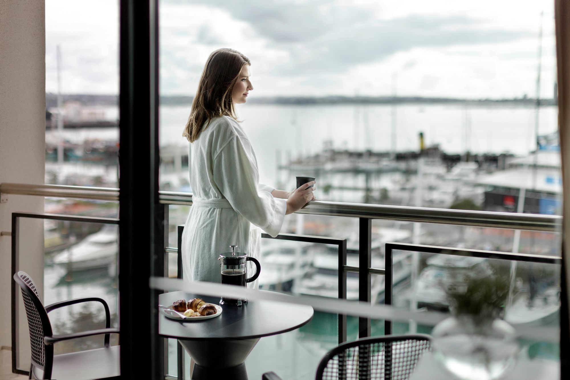 Young woman in a robe with a cup of coffee on her balcony overlooking the marina at The Sebel Auckland Viaduct Harbour