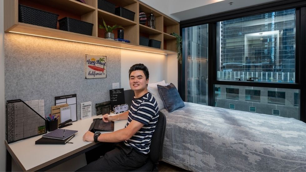 A student in a modern room with desk, bed, and city view.