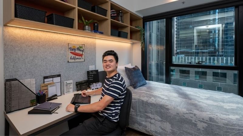 A student in a modern room with desk, bed, and city view.