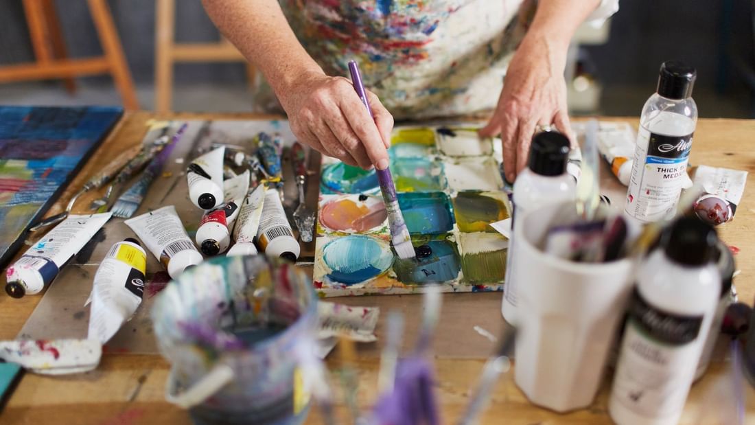 Artist painting colorful abstract artwork on canvas surrounded by paint tubes and brushes at Margaret River Open Studios