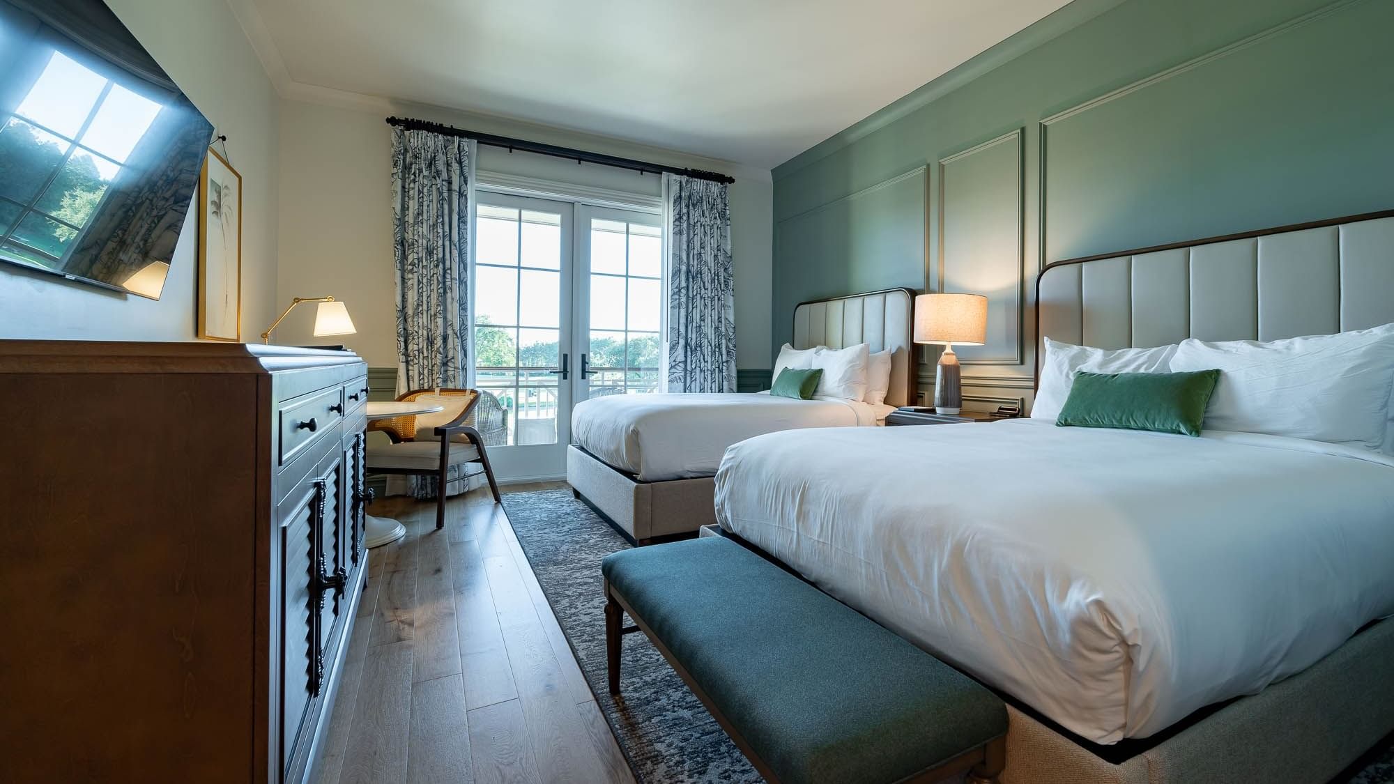 Two beds, dresser, TV, lamp, and chair in a bedroom with glass doors and green wall paneling.