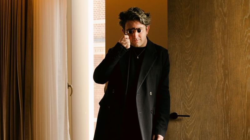 Daniel w. Fletcher in black attire standing in a Londoner bedroom, holding a pair of binoculars 