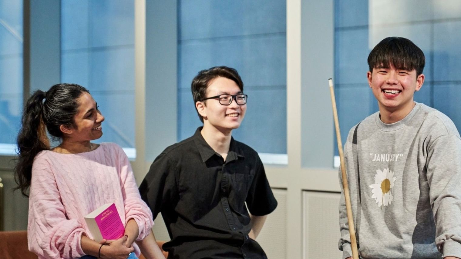 Three university students smiling and interacting by a pool table, one holding a book and another a pool stick.
