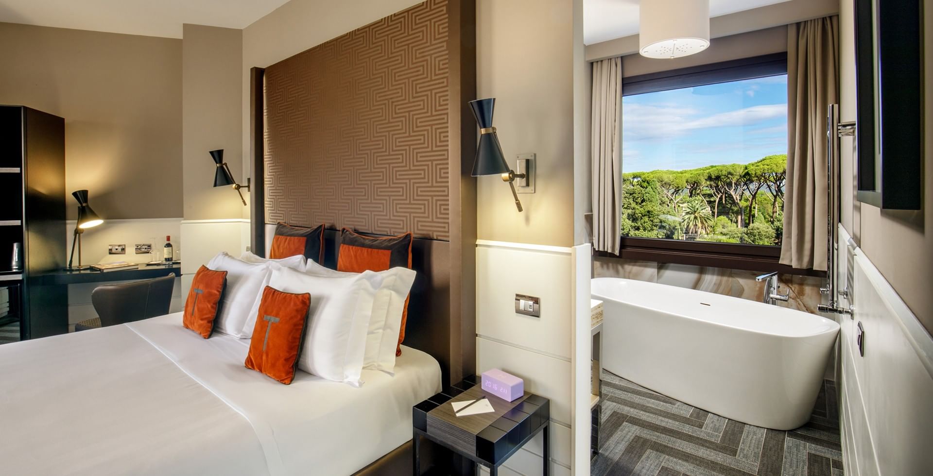 King Junior Suite Deluxe at Tribune Hotel Rome featuring a spacious king bed, modern décor with orange accents, elegant furnishings, and large windows with city and greenery views