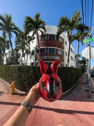 A hand holds a striking red bunny mask in front of The Gaythering surrounded by palm trees