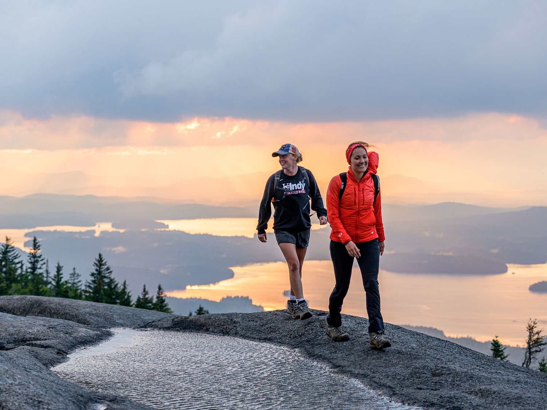 Two hikers with backpacks walk along a rocky mountain peak overlooking a sunset-lit valley near High Peaks Resort
