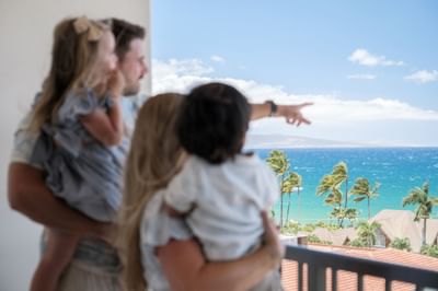 Family with two kids standing on a balcony, admiring the clear blue ocean and palm trees at the Maui Coast Hotel