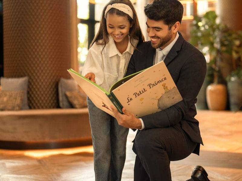 Man and girl reading a Le Petit Prince book together in hotel lobby. 