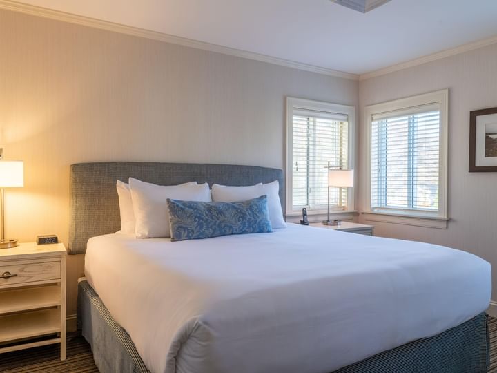 A neatly made bed with white linens and blue pillows in a room with beige walls and two windows.