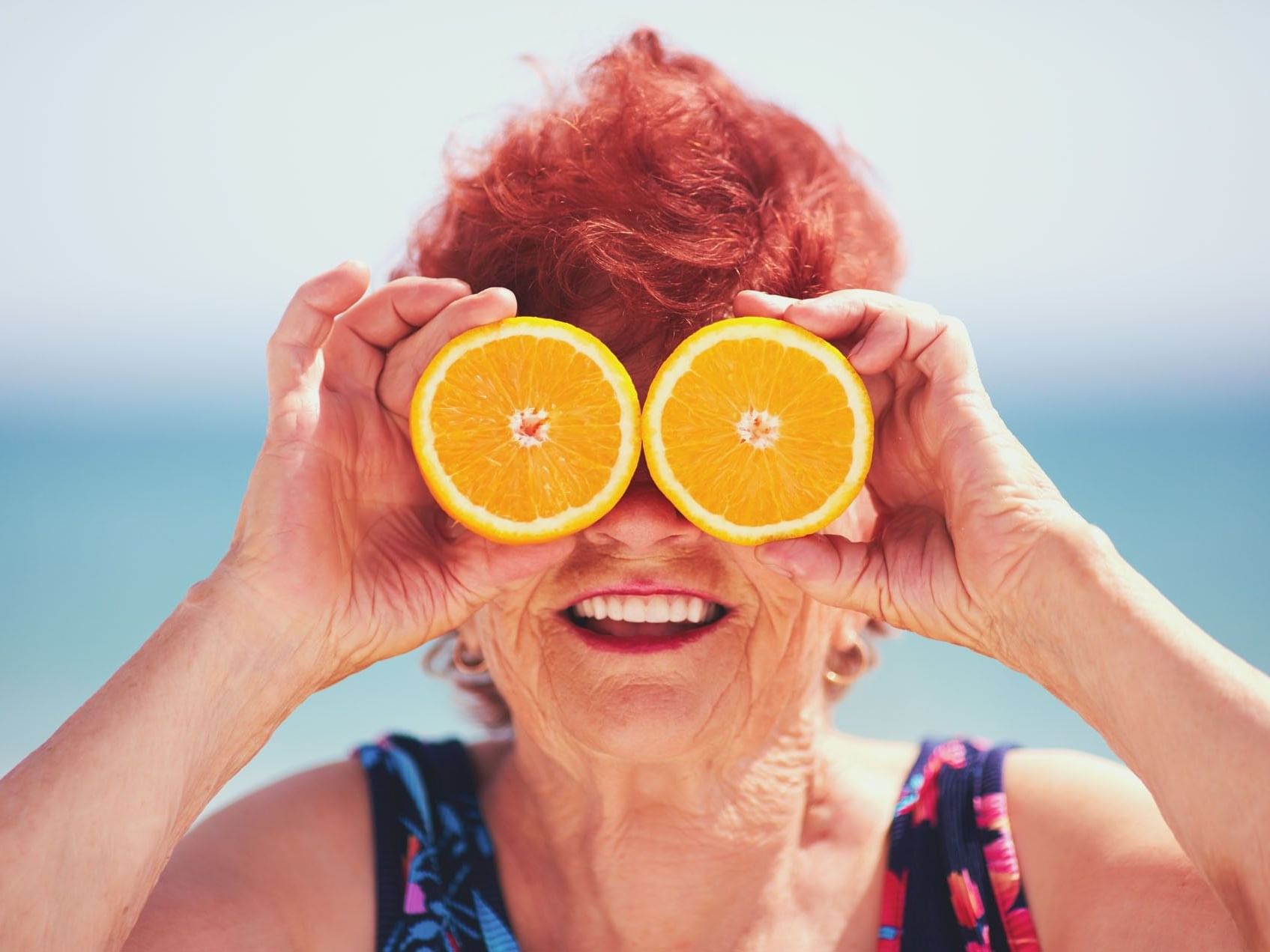 Joyful older woman with red hair holding two orange slices over her eyes at the beach near Tradewinds Apartment Hotel