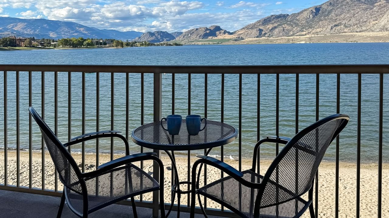 Balcony view with two chairs and coffee mugs overlooking a lake and mountains.
