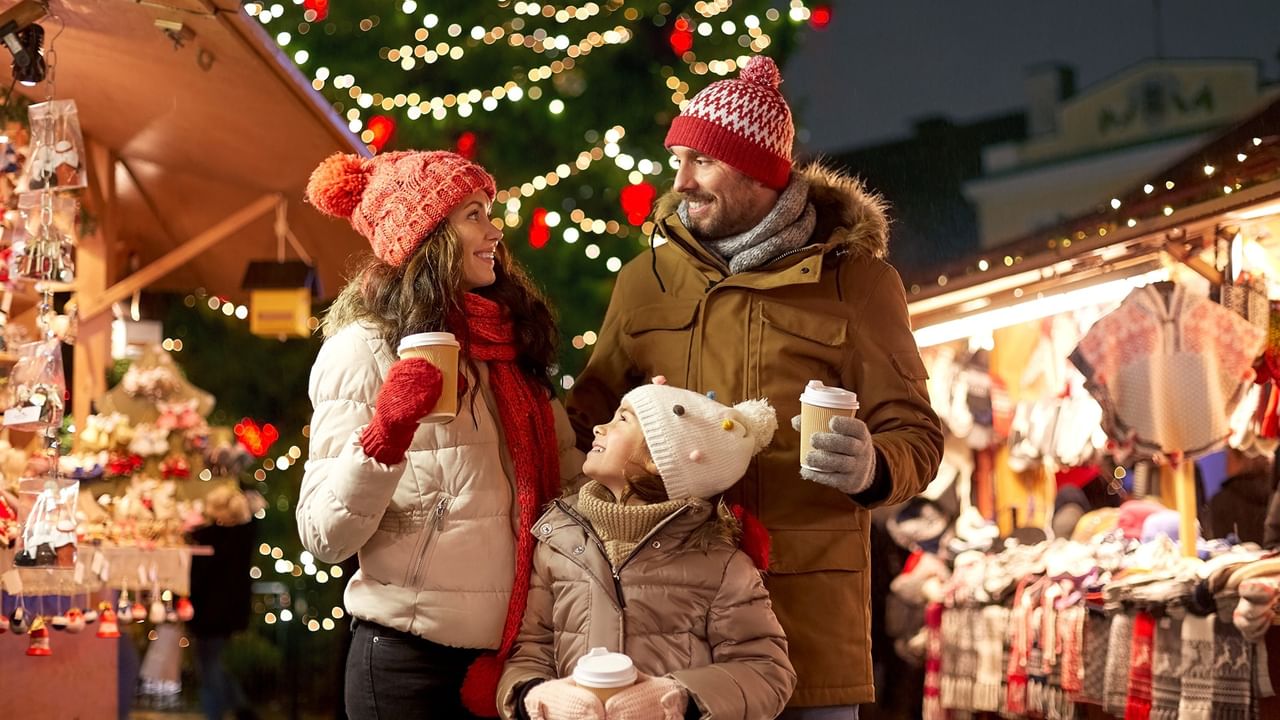 Family of three in winter clothing at a holiday market, holding coffee cups and smiling.