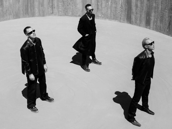 Three men in black coats and sunglasses stand in a circular concrete space.