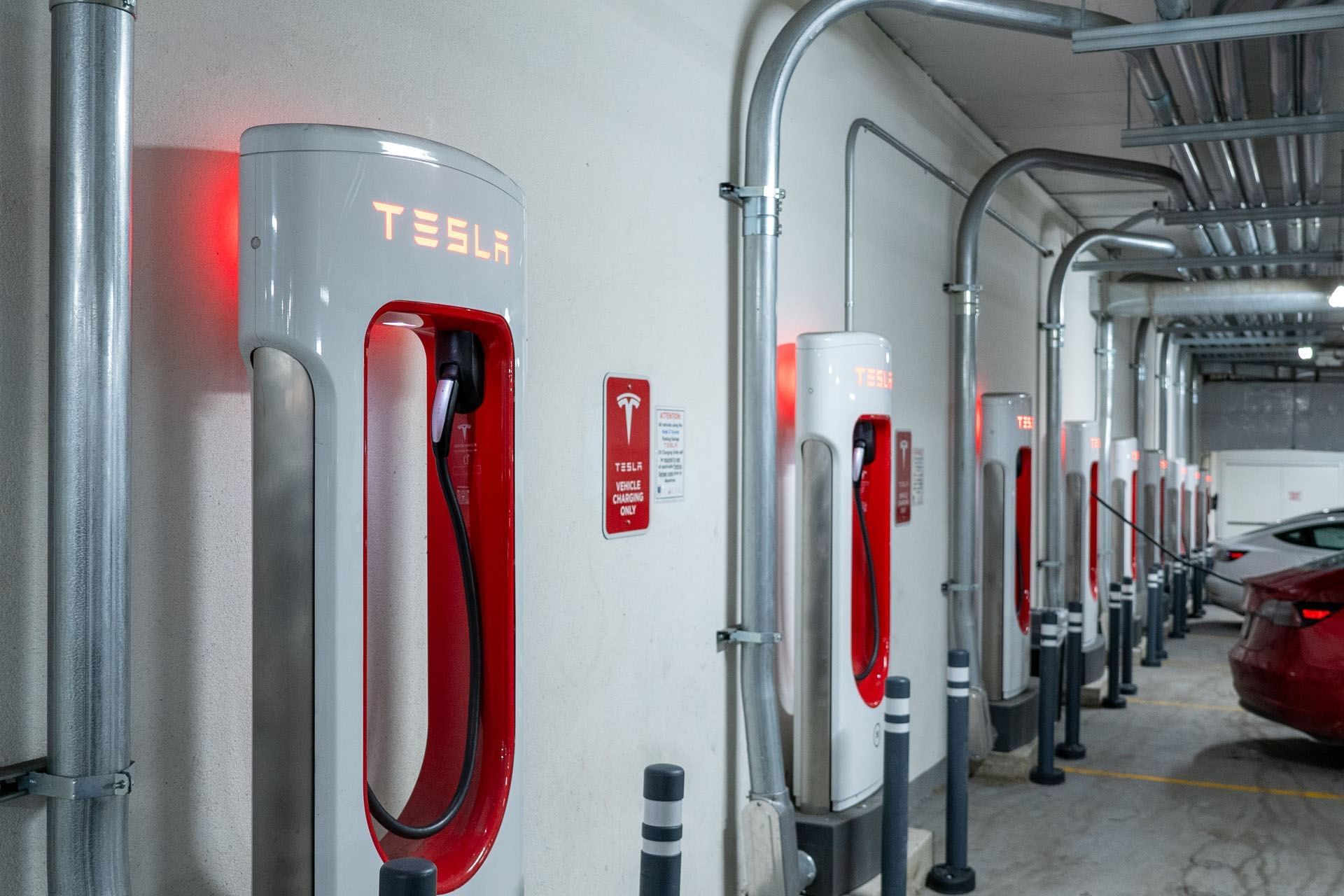 Tesla Supercharger stations lined along a covered parking area with overhead pipes and black bollards at Hotel X Toronto