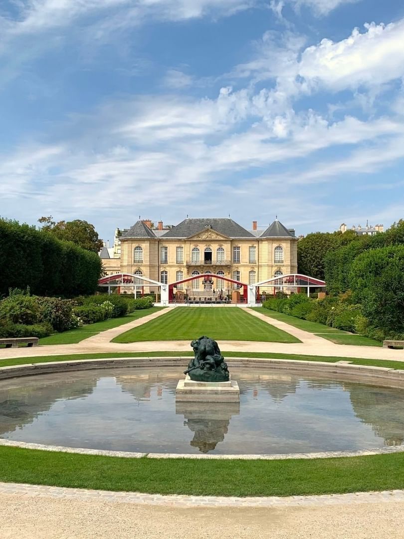 Green lawn surrounded by trees, leading to Musée Rodin by a circular pond with a sculpture near Hotel Westminster Paris