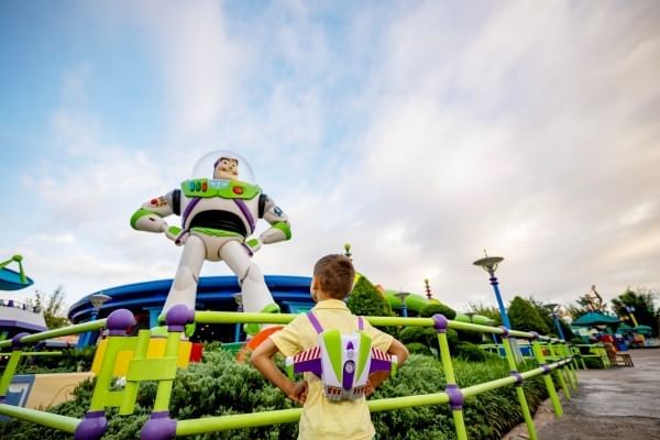 A young boy wearing a Buzz Lightyear backpack looks up at a Buzz Lightyear statue with his hands on his hips. 