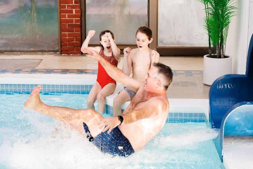 Excited father and two kids splashing into an indoor swimming pool from a blue water slide at Rodd Royalty