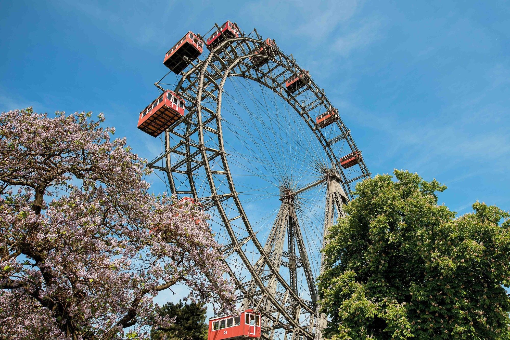 Historisches Wiener Riesenrad mit roten Gondeln vor blauem Himmel und blühenden Bäumen im Prater