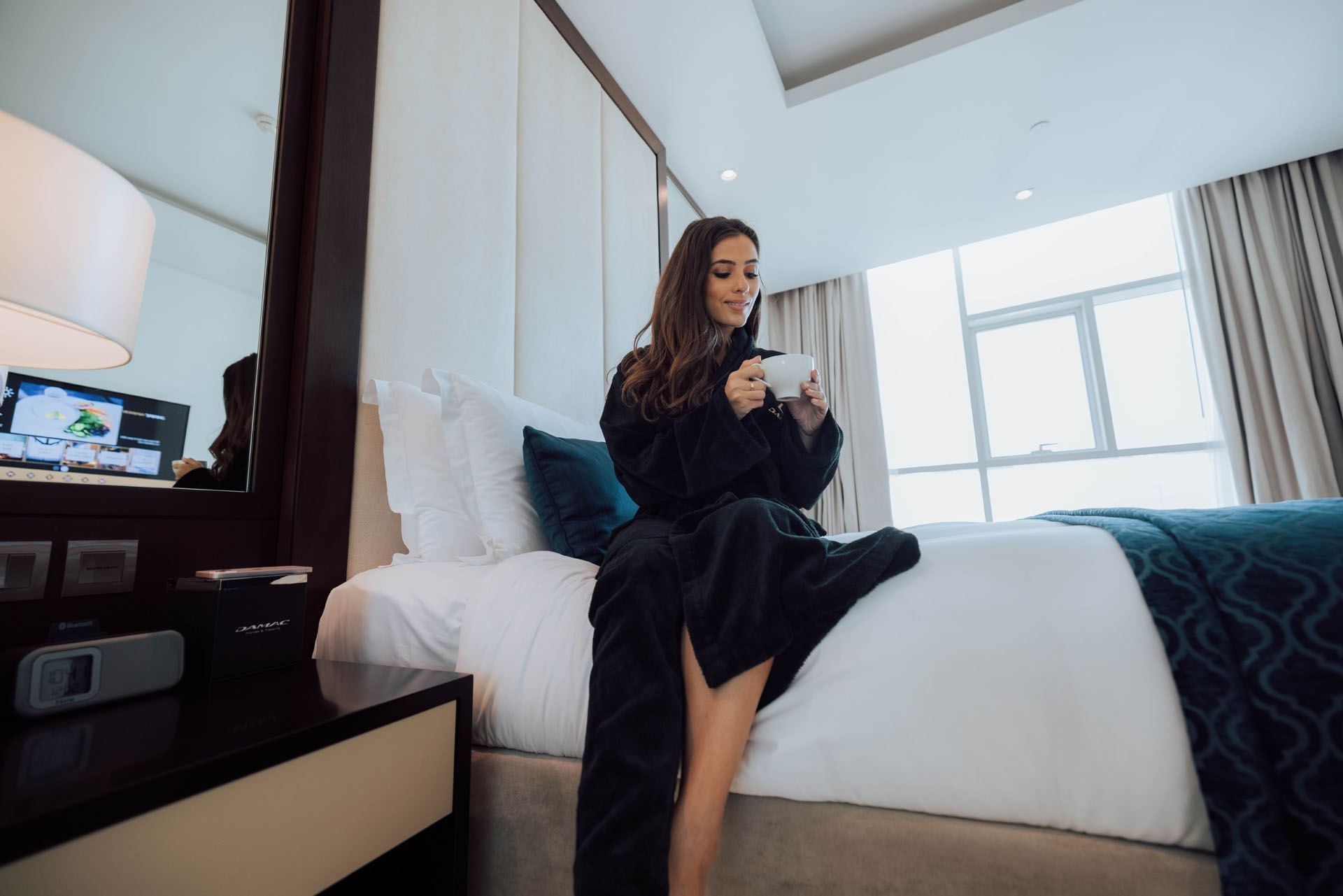 Lady sitting on bed with a cup of tea on hand in one of best dubai hotel suites at DAMAC Maison Distinction