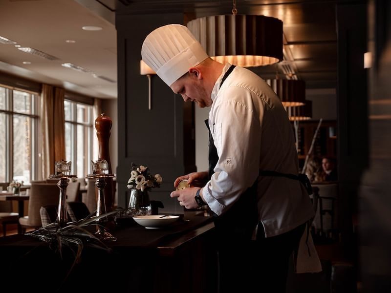 Chef in white uniform and toque preparing a dish at a restaurant counter with dim lighting.