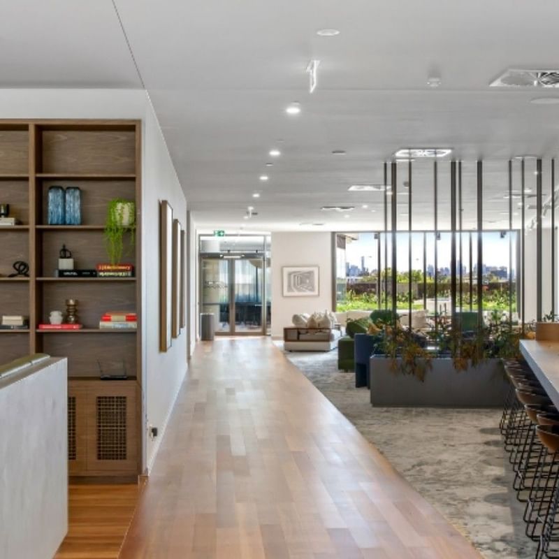 Modern communal hallway with wooden floor, bookshelves, lounge area, and large windows with city view.