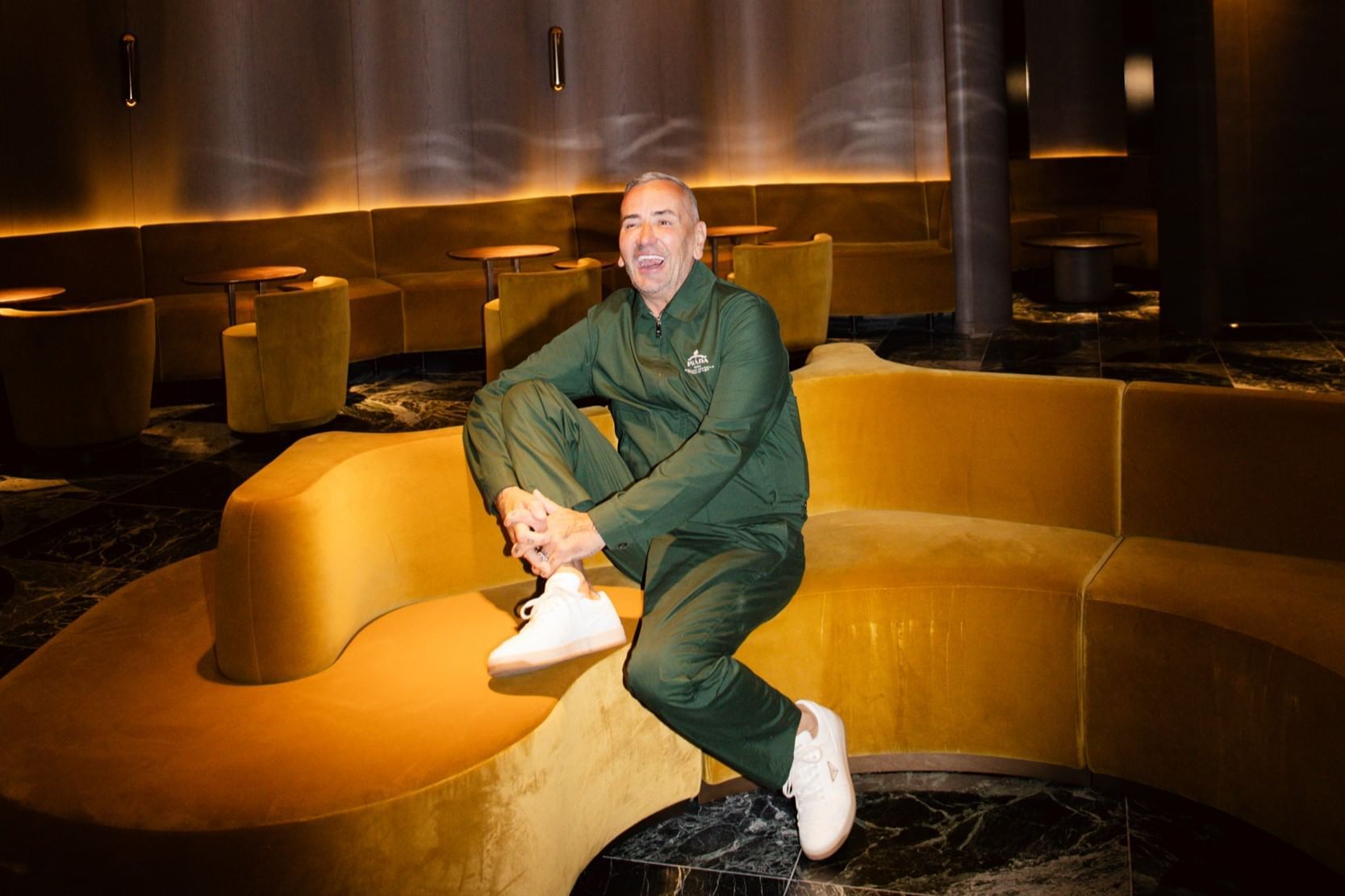 A man in a green tracksuit smiles, seated casually on a plush gold velvet sofa inside The Londoner Hotel
