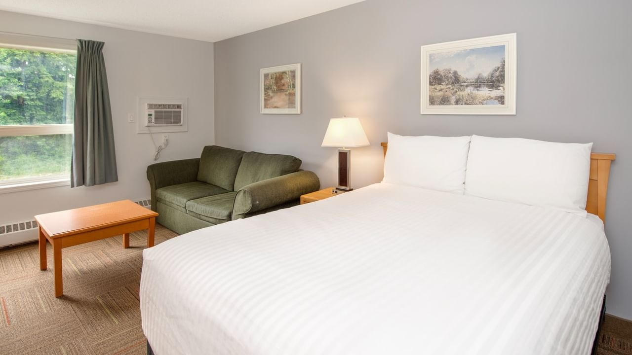 A hotel room with a white bed, green couch, window, two framed pictures, and a lamp.