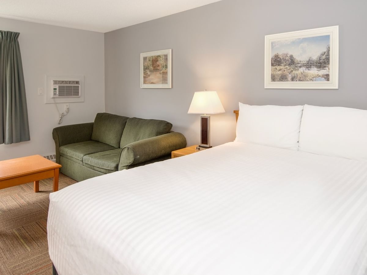 A cozy hotel room with a bed, couch, table, lamp, window, and two framed artworks.