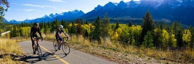 Two cyclists riding along a paved path surrounded by autumn trees and majestic mountains near Blackstone Mountain Lodge