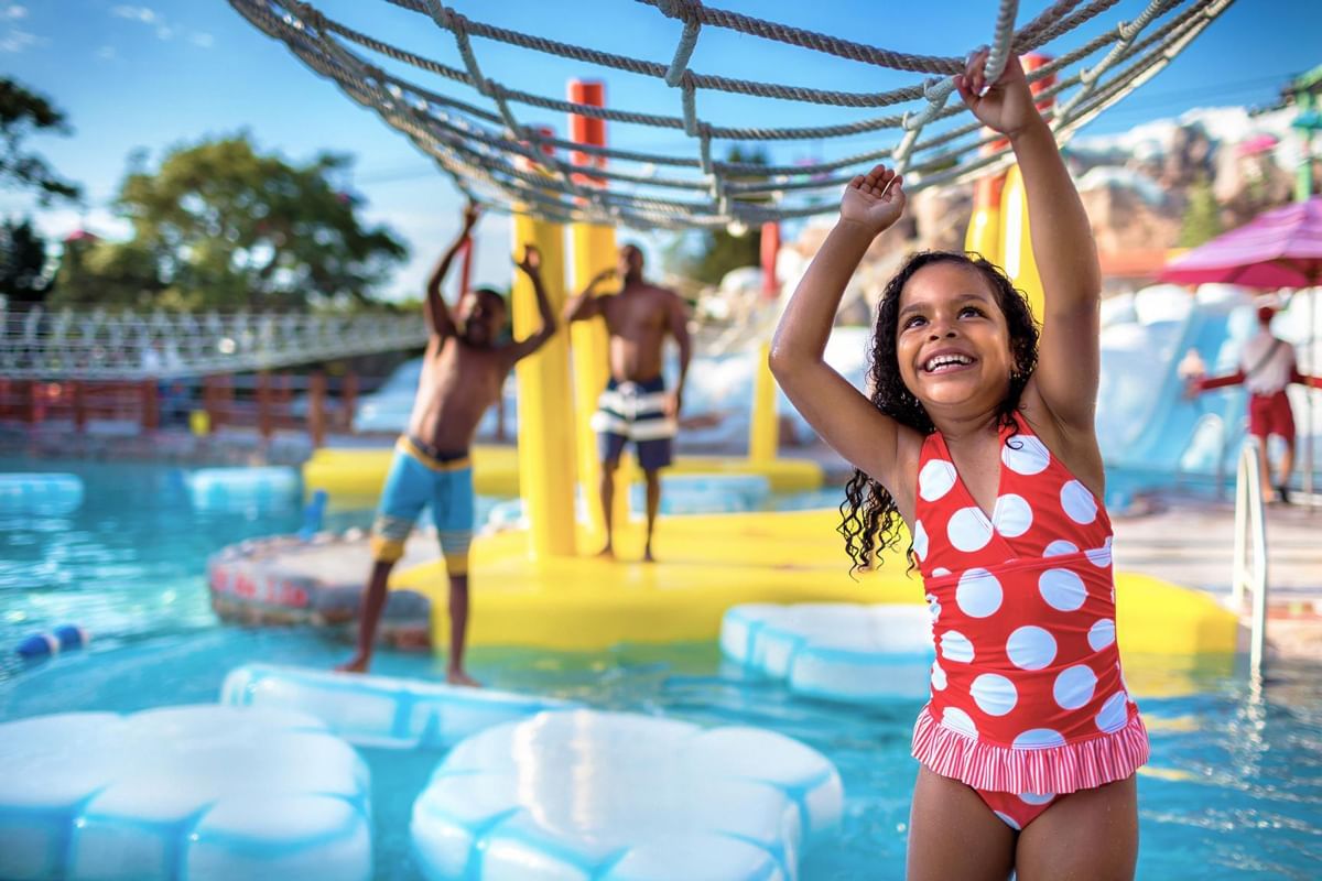 A young girl playing on a rope course in a water park with inflatable rafts and swimmers in the background.