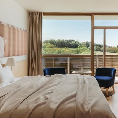 Camera da letto con letto, due poltrone blu, balcone e vista su prati e alberi.