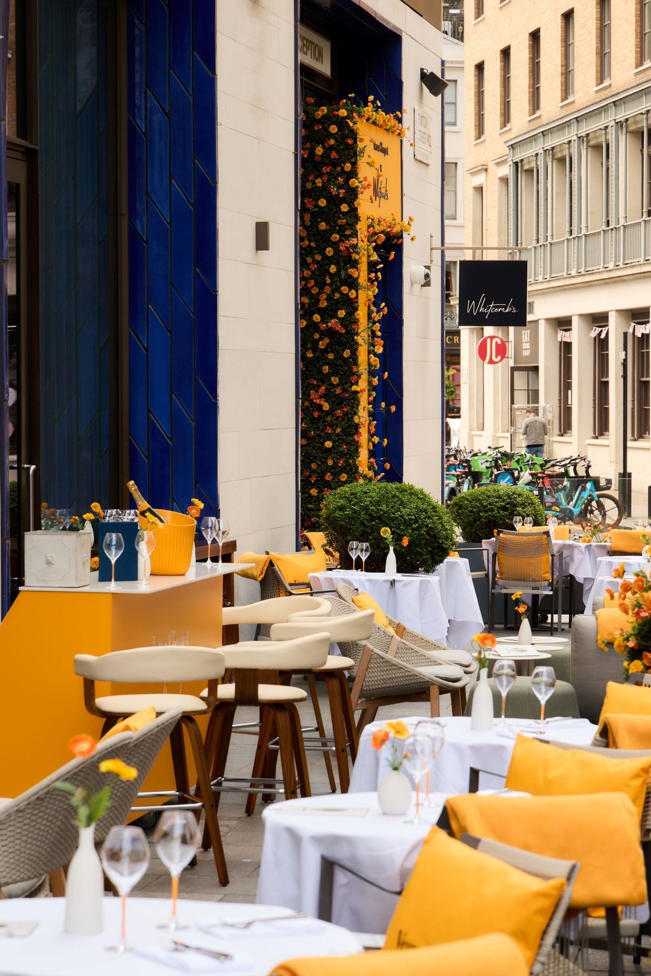 Outdoor terrace dining area with yellow and white seating, floral wall display, and champagne service at The Londoner