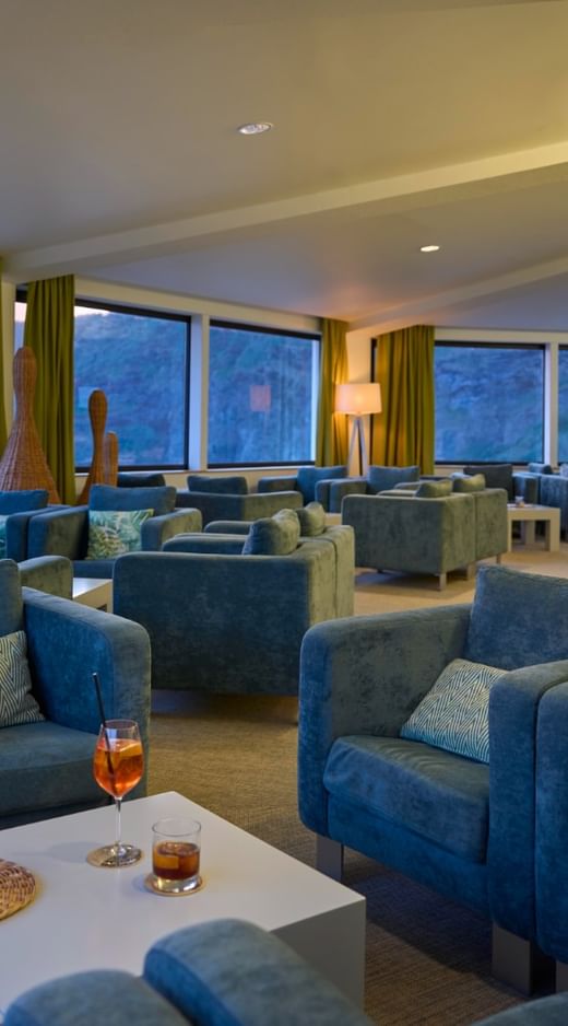 Lounge area with blue armchairs and drinks on a table facing large windows with sea views at Caloura Hotel Resort