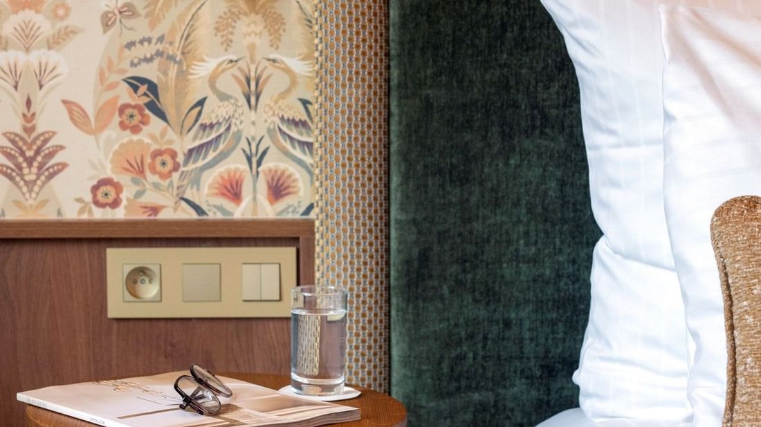 Stijlvolle hotelkamer met donkergroen fluwelen hoofdeinde, botanisch behang en gouden wandlamp naast het bed.