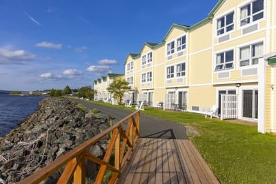 Wooden path by a yellow building under a blue sky, with a rocky shoreline at Rodd Miramichi River
