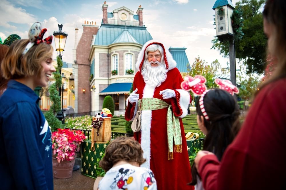 A thin man with a white beard in a red and white robe talks to two women with children in front of a French-inspired garden and building.