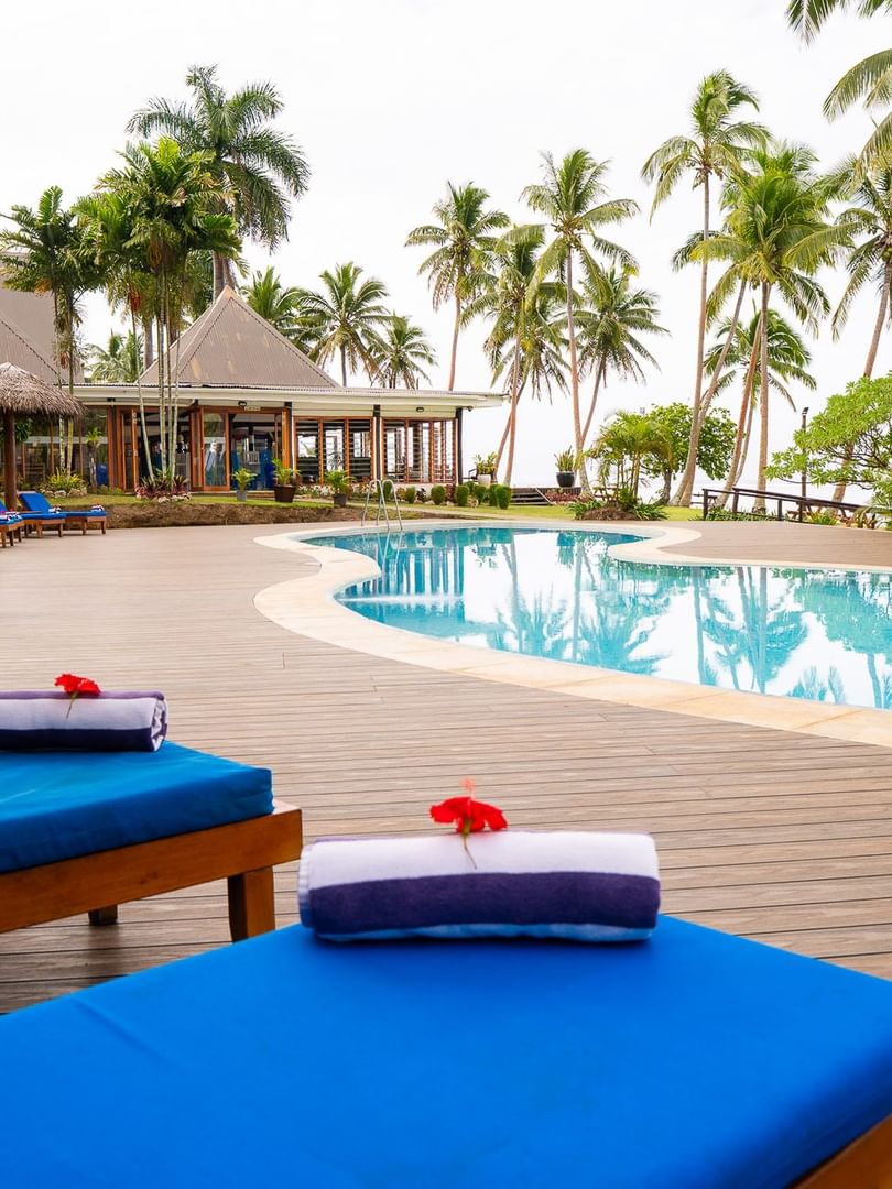 Blue cushioned loungers with white towels line a wooden deck beside a curved pool surrounded by palm trees and thatched huts.