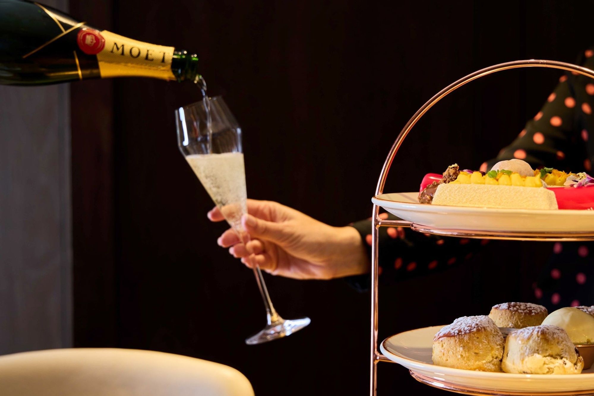 Hand pouring champagne into a glass next to a tiered plate stand with pastries and desserts.