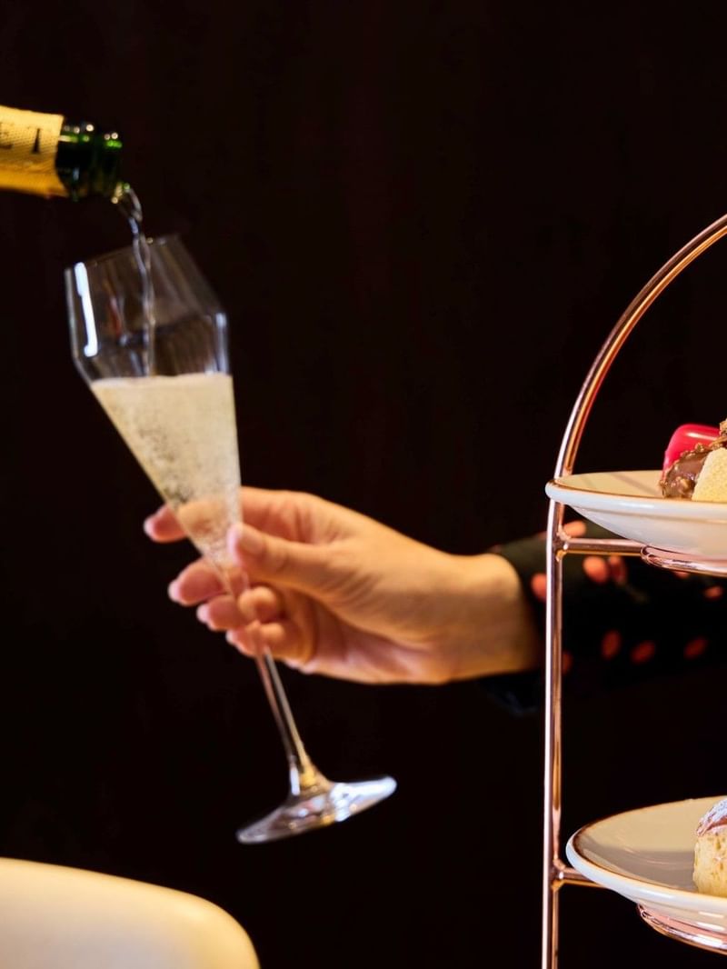 Hand pouring champagne into a glass next to a tiered plate stand with pastries and desserts.