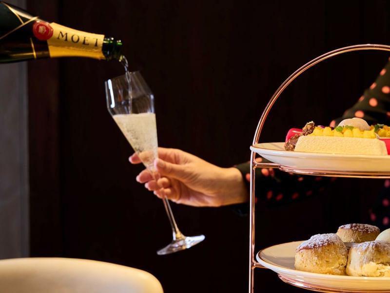 Hand pouring champagne into a glass next to a tiered plate stand with pastries and desserts.