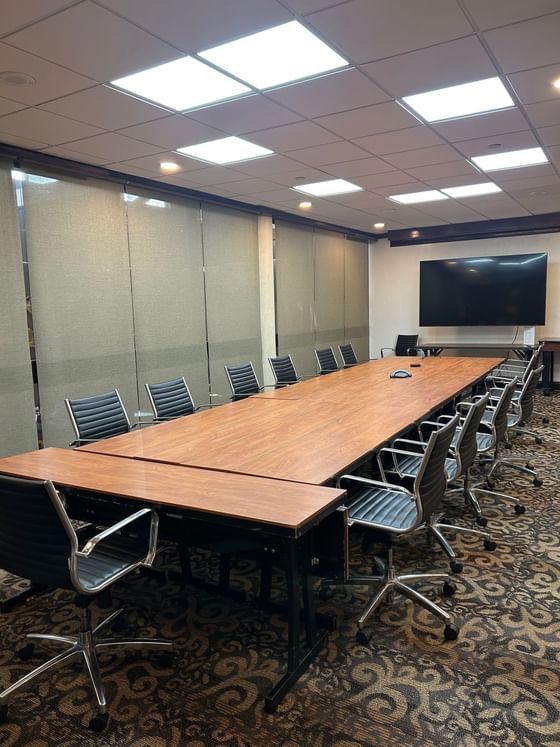 Meeting table with black office chairs facing a wall-mounted TV near window shades in The Mezzanine at Miami Lakes Hotel