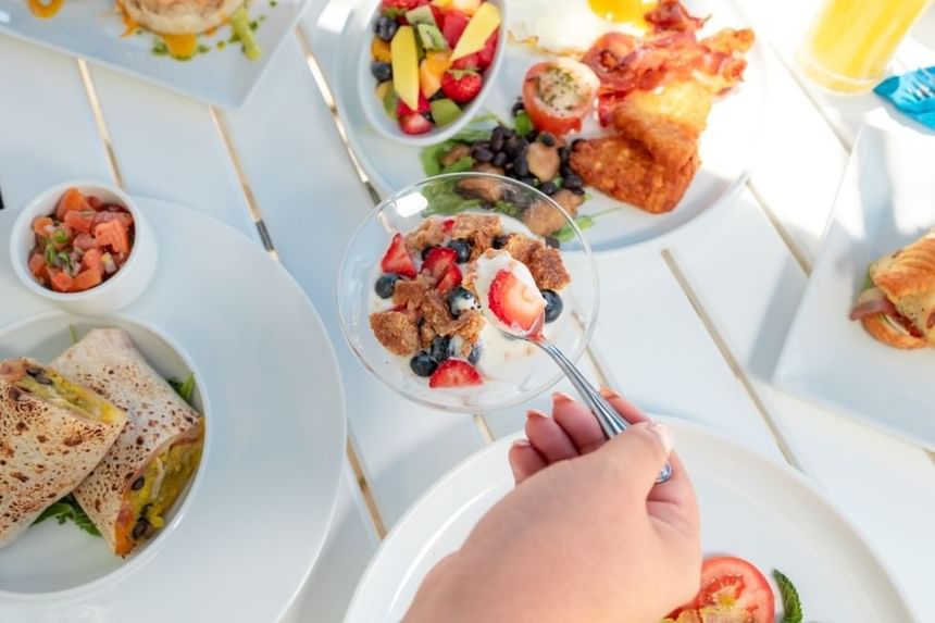 A person's hand holds a spoon over a bowl of yogurt with berries, among various dishes on a table.