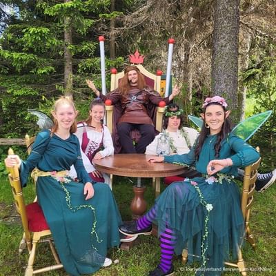 Five people in fairy costumes sit around a table, one man dressed as a king sits on a throne.